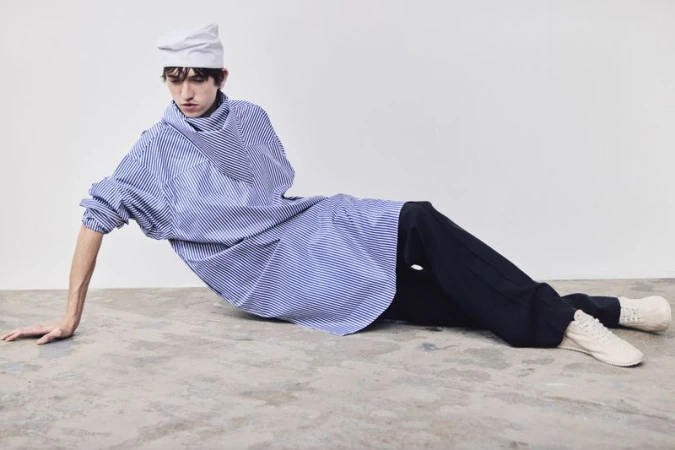 Adult male fashion model poses lying down wearing a striped shirt, dark pants, white shoes, and white cap against a white background.