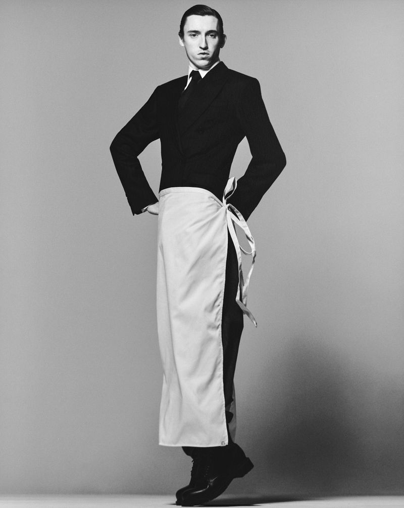 A man stands in a photo studio in black formal wear with a white apron. He poses with his hands on his hips and looks at the camera.