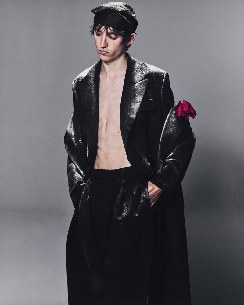 Man in a dark, shimmering jacket with a red rose, posing against a grey background in a studio fashion shot. Model wears a dark cap.