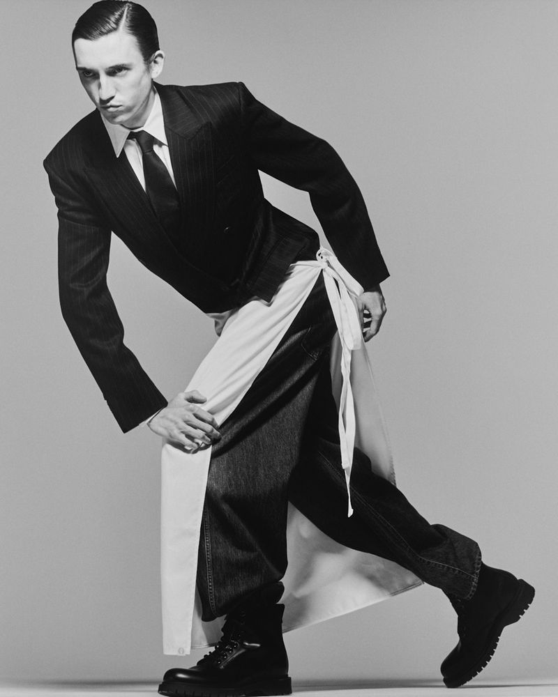 Adult male in suit and jeans poses in a studio with a white apron tied around his waist. The shot is in black and white.