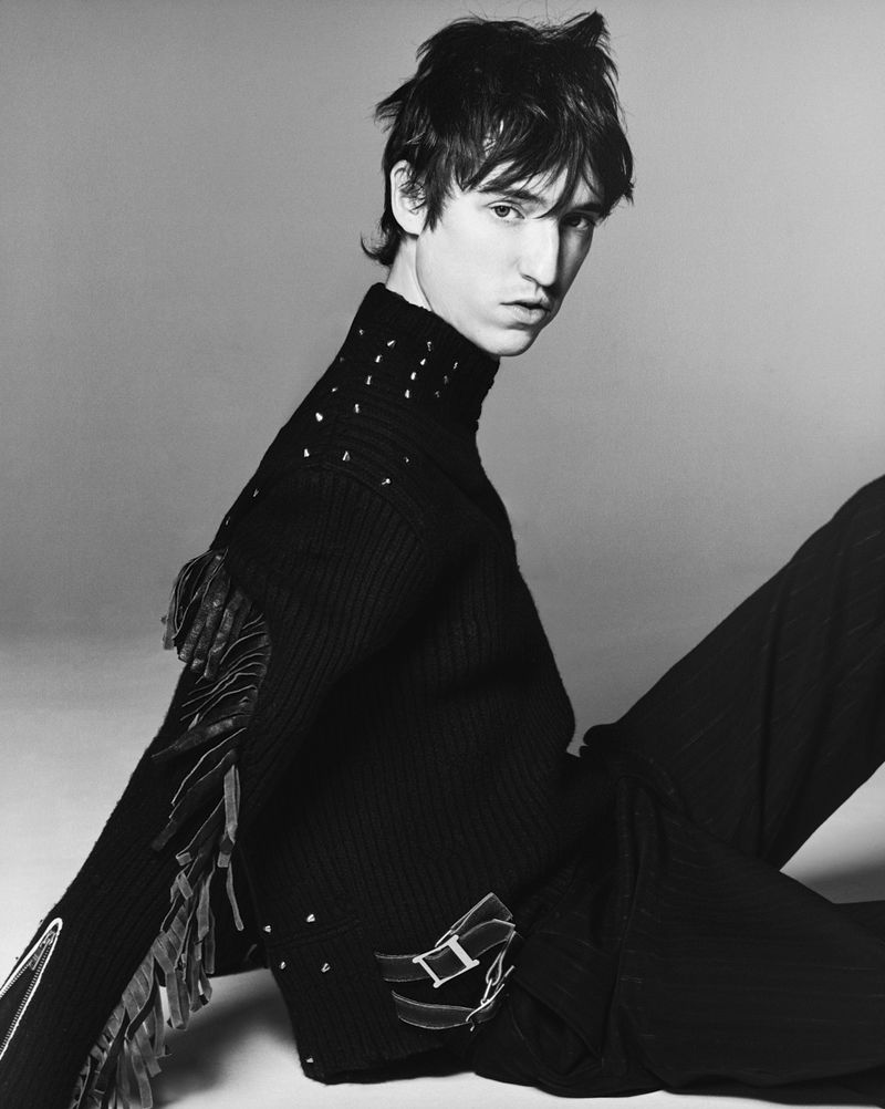 Black and white portrait of a young male model wearing a studded black turtleneck sweater with leather fringe on a grey backdrop.