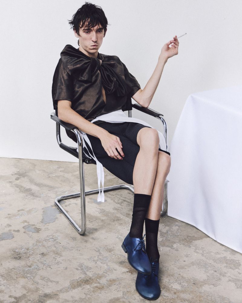 An androgynous person sits in a metal chair in a studio, dressed in an avant-garde outfit, smoking a cigarette with a white background.