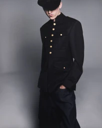 Male model wearing a black military tunic jacket with gold buttons and a black peaked cap against a grey studio backdrop.