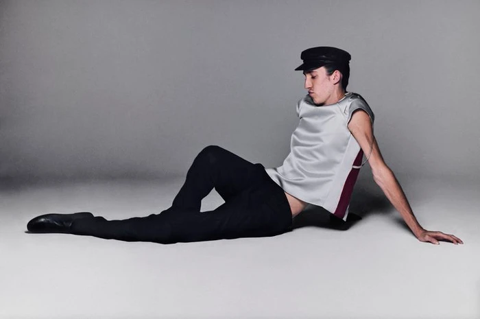 A young man wearing a black cap, silver sleeveless top, and black trousers reclining on a grey studio floor.