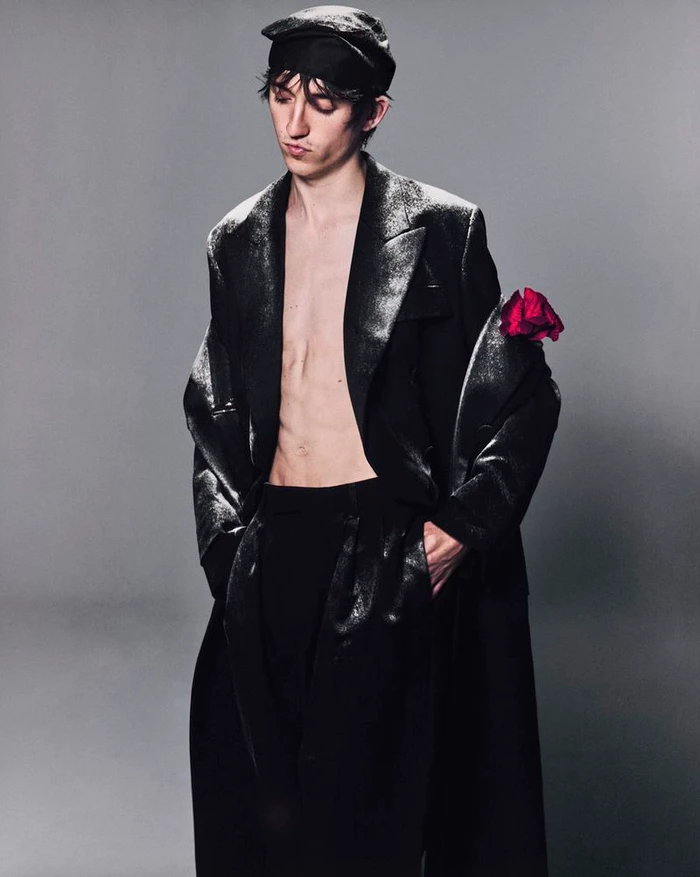 Fashion model posing shirtless in an open textured black coat and cap with a red rose against a grey studio backdrop.
