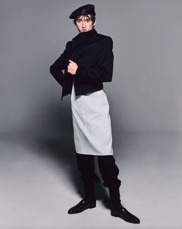 Male model posing in a black cropped jacket, white apron tunic, black trousers, and a beret against a grey backdrop.