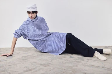 Model reclining on a studio floor wearing an oversized blue and white striped tunic, dark trousers, and white folded headwear.