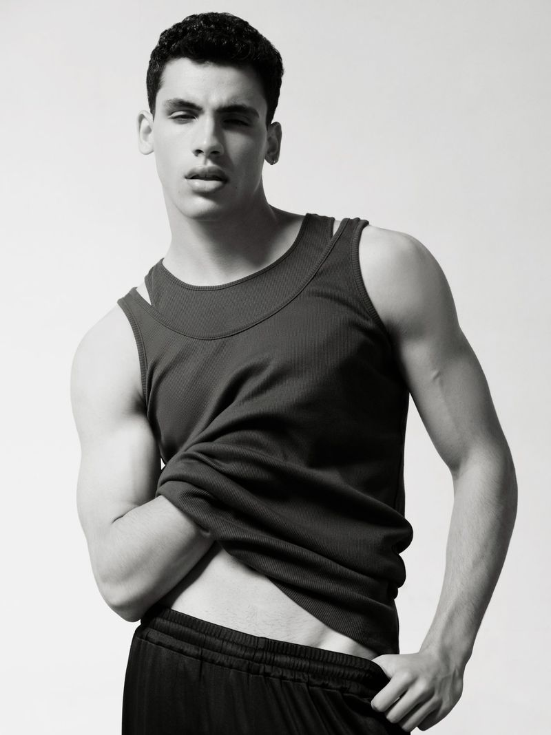 Studio shot of a young adult male in a tank top and athletic shorts, posing against a white background, with a black and white color palette.