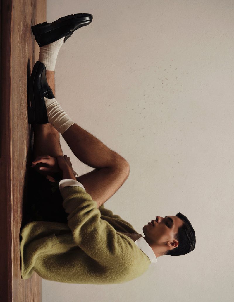 Man in an olive green cardigan, white collared shirt, and black loafers seated on a wooden floor against a neutral wall.