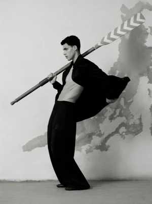 Monochrome editorial portrait of a shirtless man holding a rowing paddle across his shoulders against a textured wall.