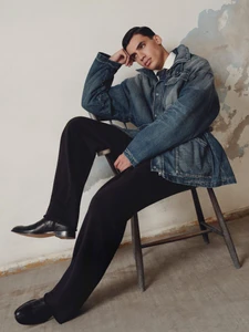 Man seated on a wooden chair wearing a denim jacket and dark trousers against a distressed wall.