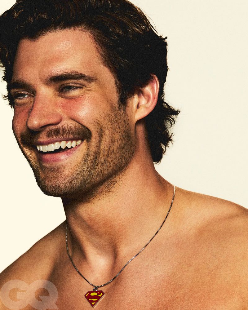 Smiling man with bare chest wearing a necklace with a Superman logo pendant against a light beige background in a close-up portrait.