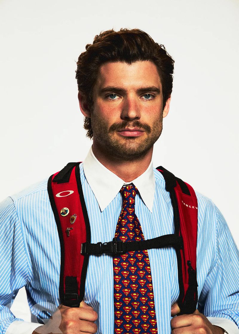 Portrait of a man in a blue striped shirt and tie with a red backpack against a white background, shot with a medium camera framing.