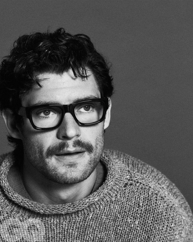 Monochrome portrait of a man with dark hair wearing black-framed glasses and a gray knitted sweater against a gray backdrop.