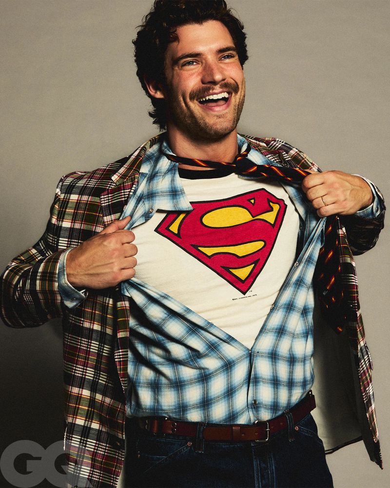 Enthusiastic adult male revealing a Superman t-shirt beneath a plaid blazer and blue checked shirt indoors against a grey backdrop.
