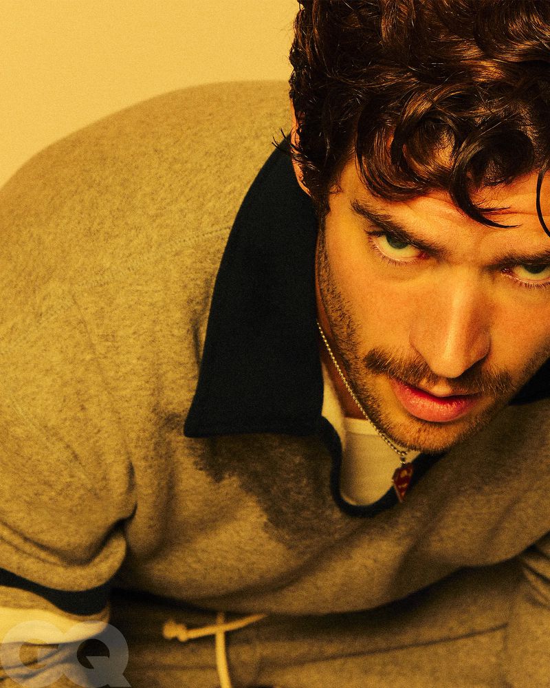 Portrait of an adult male model wearing a gray sweatshirt with curly brown hair, posing indoors in front of a golden yellow background.