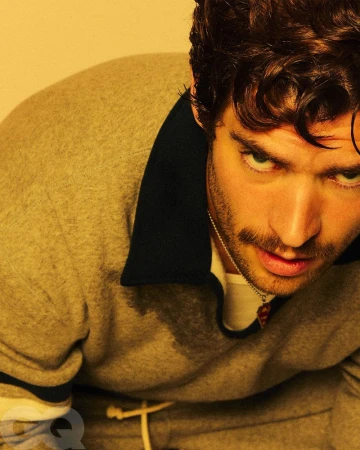 Portrait of an adult male model wearing a gray sweatshirt with curly brown hair, posing indoors in front of a golden yellow background.