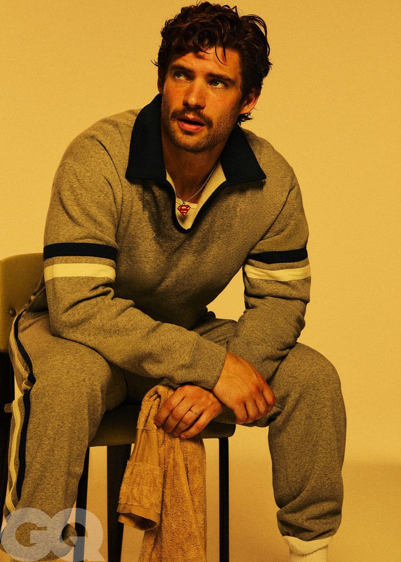 A tanned adult male with dark curly hair and a mustache poses indoors in a gray sweat suit, seated on a chair with a towel.