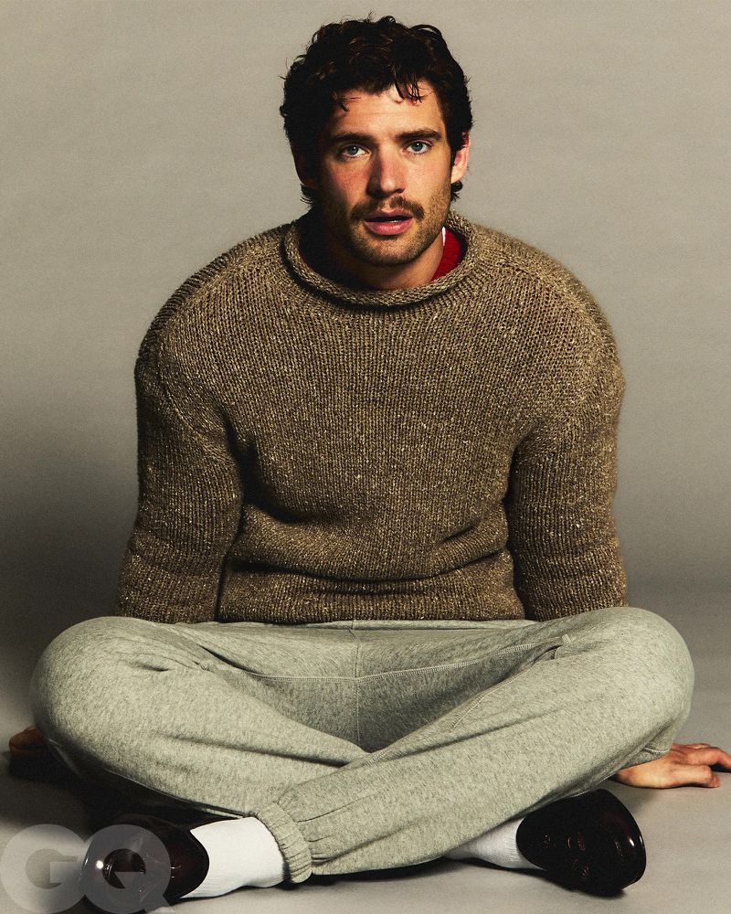 Portrait of a dark-haired adult male with a mustache wearing a sweater and sweatpants sitting cross-legged in a photo studio.
