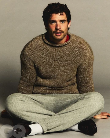 Portrait of a dark-haired adult male with a mustache wearing a sweater and sweatpants sitting cross-legged in a photo studio.