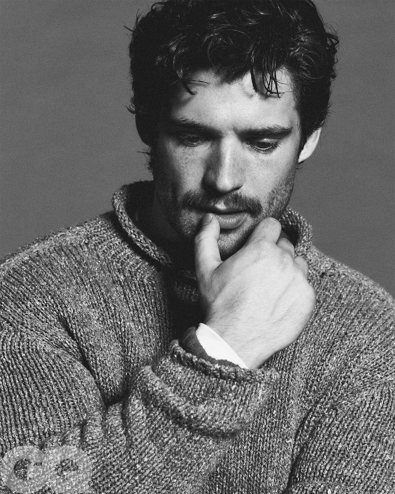 Black and white portrait of a man with a mustache wearing a knit sweater, his hand thoughtfully placed near his face.