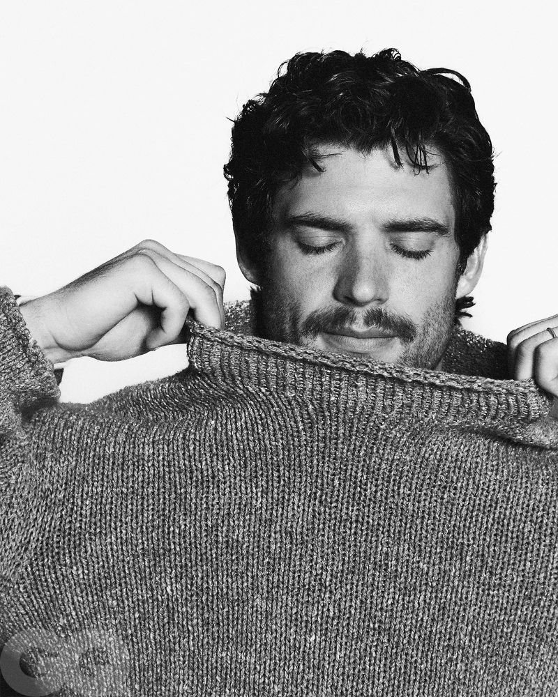 Black and white portrait of a male model pulling a gray wool sweater up to his face in a photo studio with natural lighting.