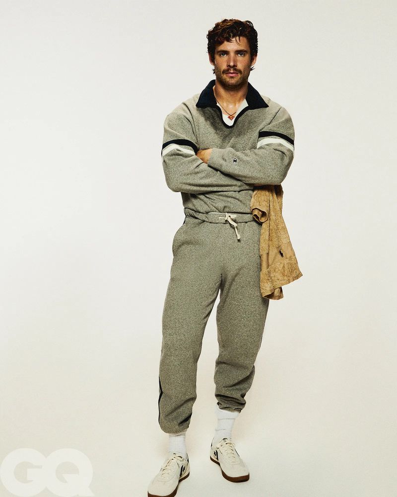 An adult male in a gray tracksuit poses in a studio with arms crossed, wearing white sneakers, against a plain white background.