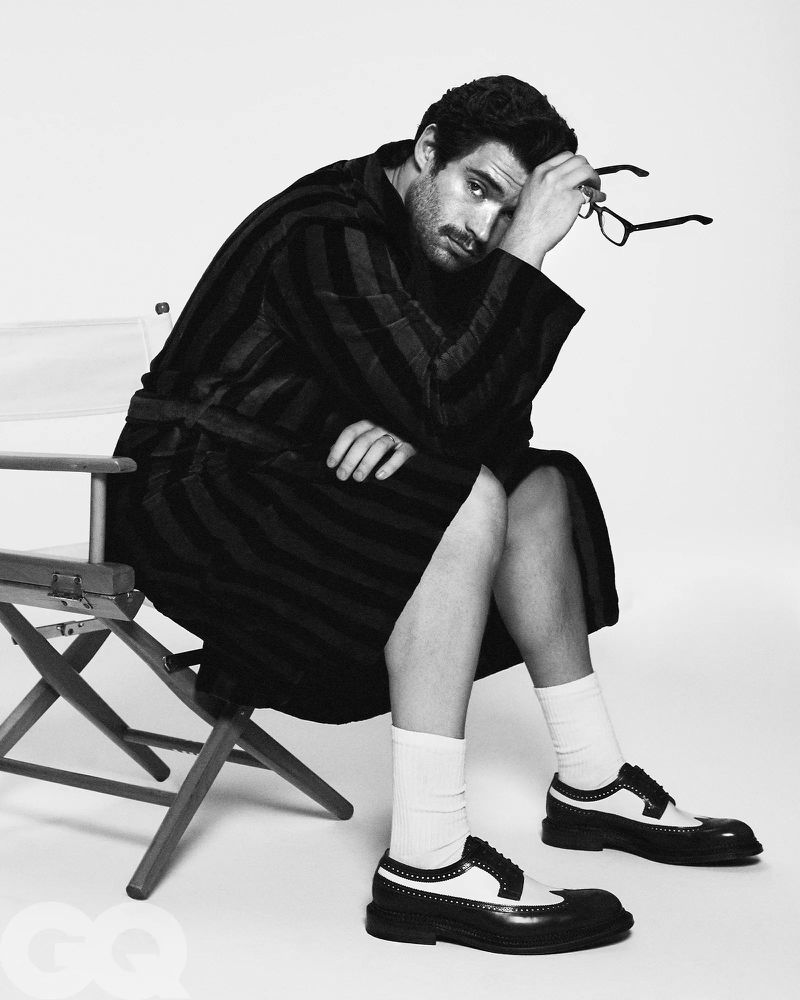 Monochrome portrait of a man seated on a director's chair wearing a striped robe and dress shoes, holding glasses in studio lighting.