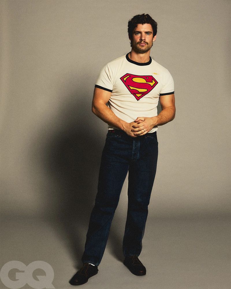 A fair-skinned man poses in dark blue jeans and a Superman T-shirt against a gray backdrop, lit with photo studio lighting.