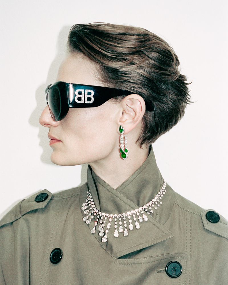 A fair-skinned adult female poses in a studio wearing a trench coat, sunglasses with a logo, a diamond necklace, and dangling earrings.