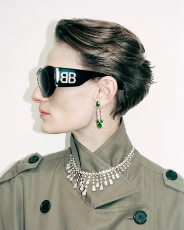 A fair-skinned adult female poses in a studio wearing a trench coat, sunglasses with a logo, a diamond necklace, and dangling earrings.