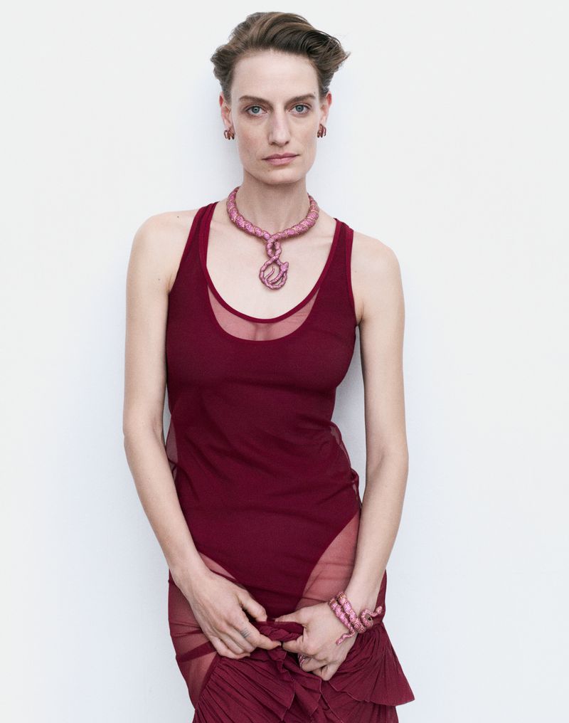 Adult woman in a maroon dress with sheer skirt panels stands against a white wall, adorned with a pink necklace and bracelet.