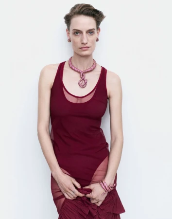 Adult woman in a maroon dress with sheer skirt panels stands against a white wall, adorned with a pink necklace and bracelet.