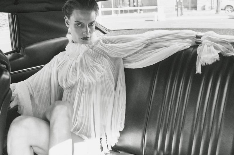 Monochrome fashion portrait of a model in a pleated white dress with dramatic sleeves, seated in the back of a luxury car.