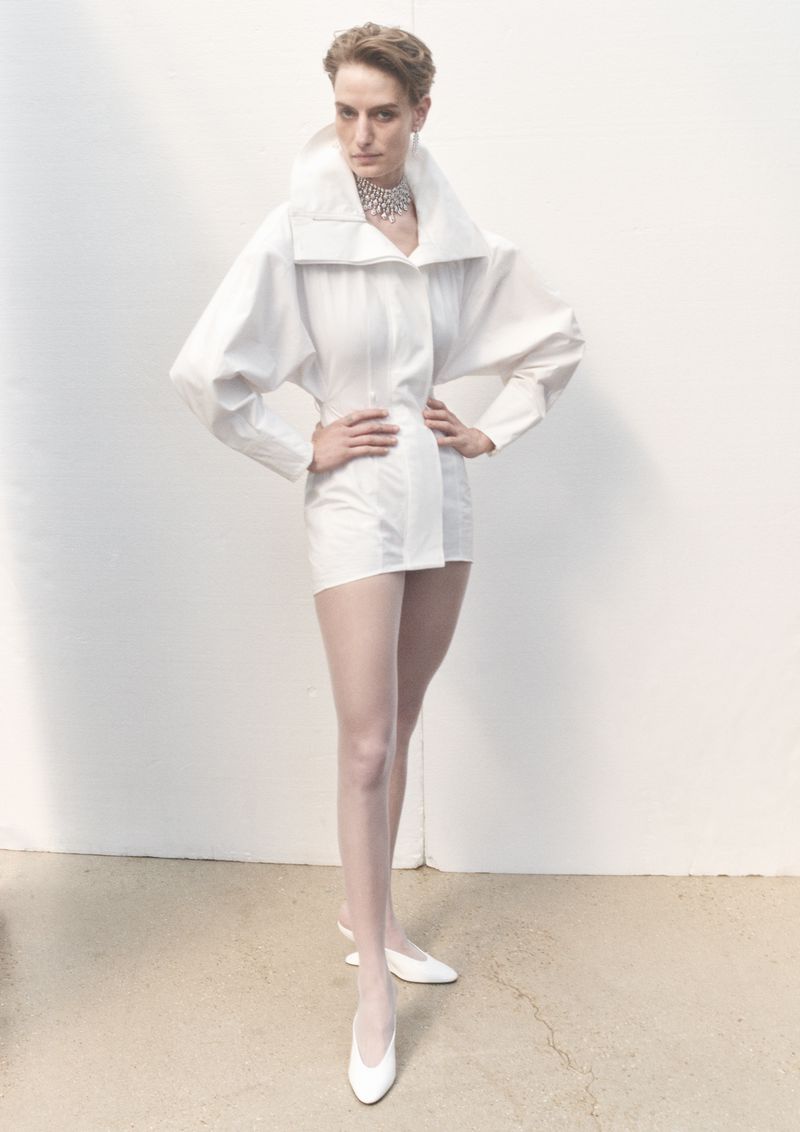 A fashion shot of a woman posing in a white, oversized shirt dress with white tights and heels in front of a white wall.