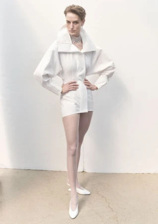 A fashion shot of a woman posing in a white, oversized shirt dress with white tights and heels in front of a white wall.