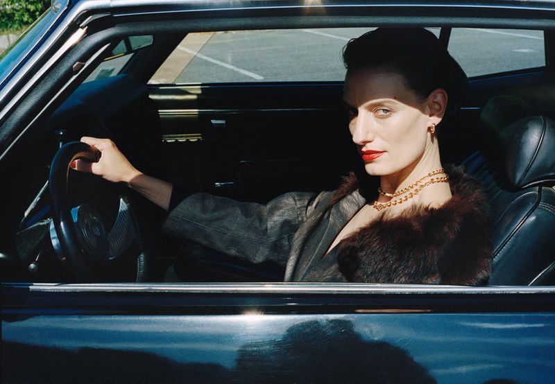 Model Veronika Kunz wearing a fur-collared coat and gold necklace sitting behind the steering wheel of a vintage car.