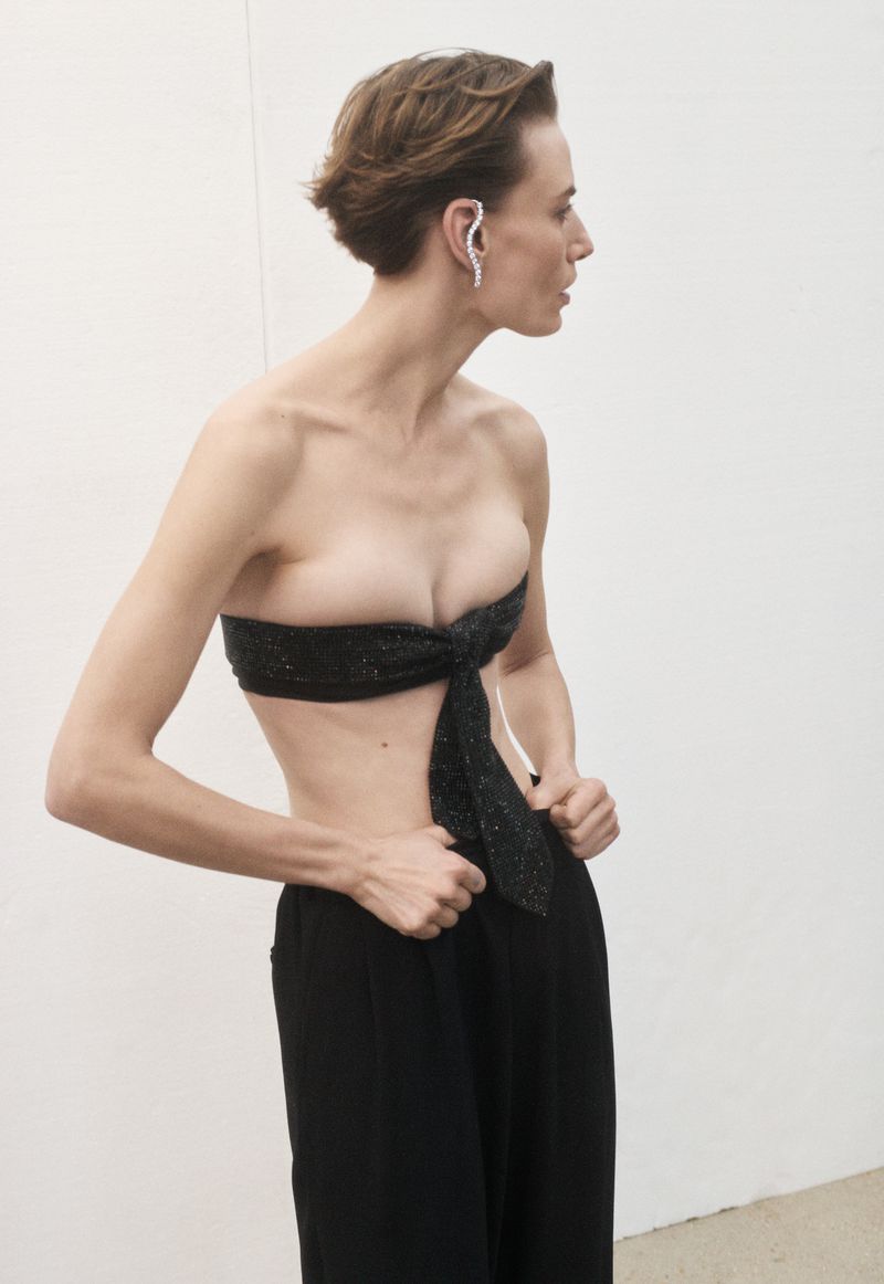 Adult female fashion model poses in a tied black bandeau top and black pants against a white wall, looking off to the side.