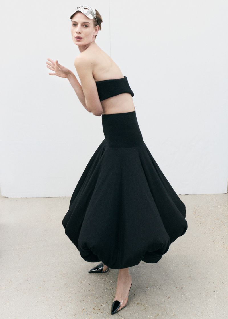 Adult female fashion model poses in a black crop top and voluminous black skirt in a studio with white walls, silver eye mask and black heels.