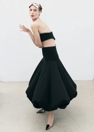Adult female fashion model poses in a black crop top and voluminous black skirt in a studio with white walls, silver eye mask and black heels.