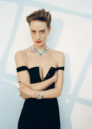 An adult female model poses in an off-the-shoulder black dress, wearing statement jewelry and bold red lipstick, against a neutral backdrop.