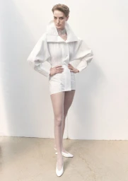 Model Veronika Kunz wearing a high-collar white structural jacket dress with white heels against a pale backdrop.
