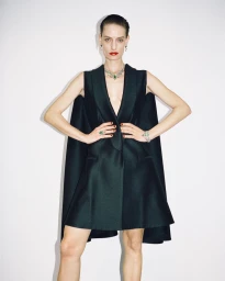 Model Veronika Kunz wearing a dark green sleeveless dress with a cape back, emerald jewelry, and red lipstick against a white background.