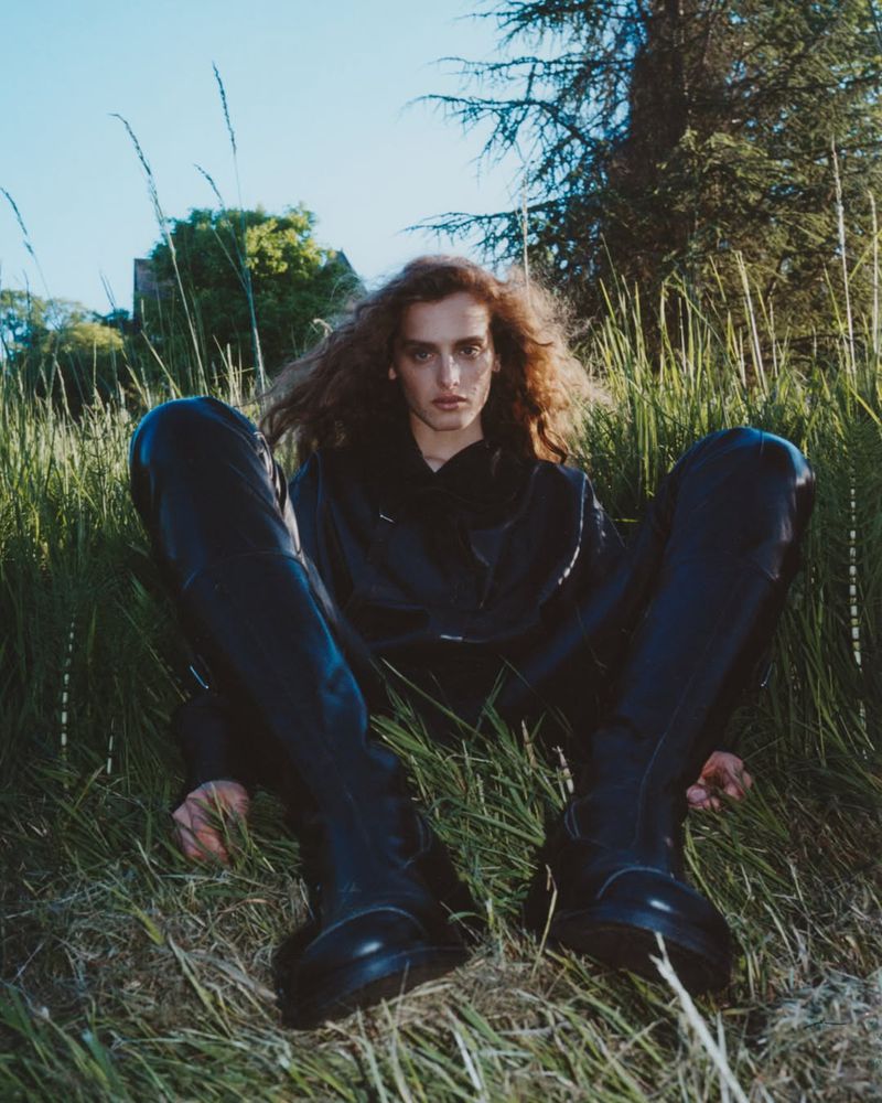 Model with long wavy hair seated in tall grass wearing an all-black outfit with prominent chunky knee-high leather boots.