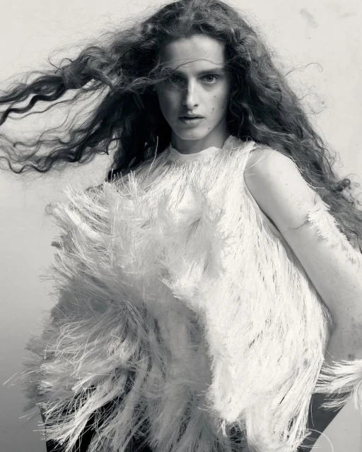 Monochrome portrait of a young woman with curly hair wearing a fringed top against a light background, looking directly at the camera.