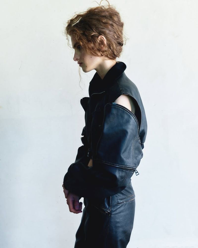 A slender person with curly hair in black leather jacket and pants poses against a light backdrop in a fashion-forward editorial shot.