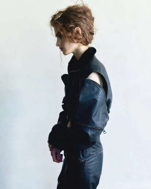 A slender person with curly hair in black leather jacket and pants poses against a light backdrop in a fashion-forward editorial shot.