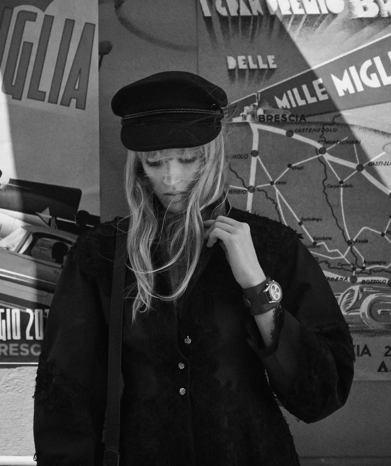 Blonde woman in a black jacket and cap poses in front of an old Italian travel poster in a monochromatic fashion editorial image.