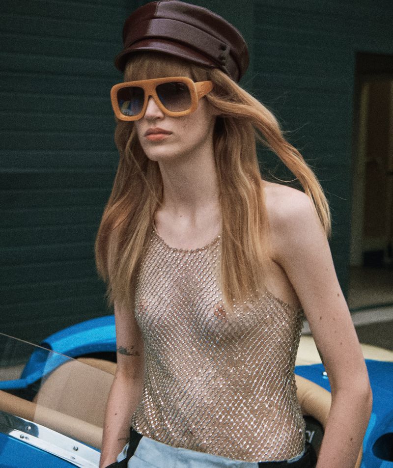Fashionable adult woman wearing a brown hat and sunglasses with a vintage car in the background, striking a pose in an outdoor setting.