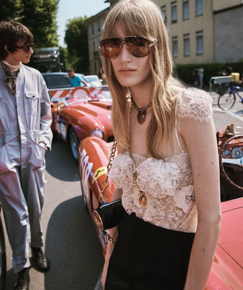 Fashion shot featuring a woman in a lace top and sunglasses beside a red vintage race car, with a man in a jumpsuit in the background.
